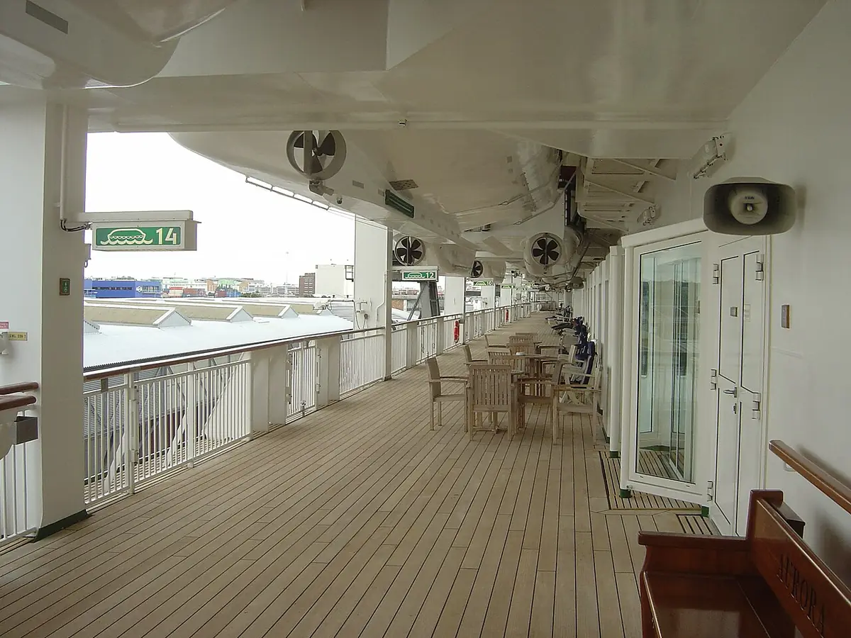 Convés de passeio de um navio de cruzeiro, com piso de madeira e cadeiras ao longo da amurada